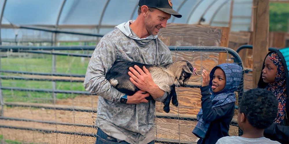 Man holding goat while children interact joyfully inside greenhouse farm